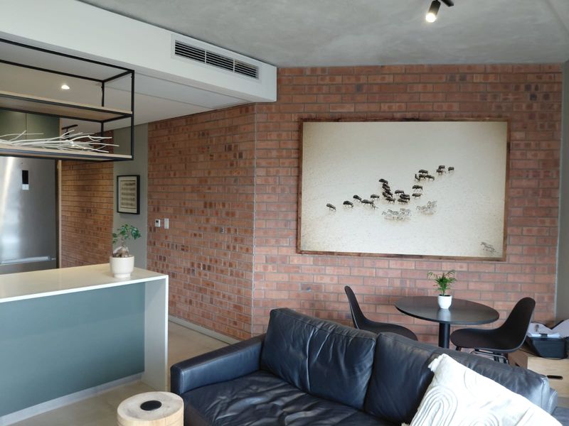 Modern open-plan living area featuring an exposed brick feature wall, concrete ceiling, ducted air conditioning, and a sleek kitchen island.