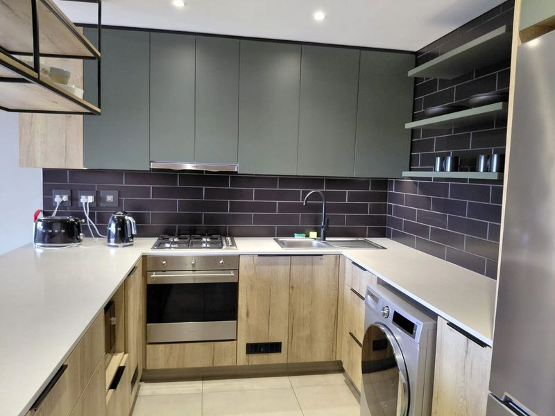 Modern kitchen featuring matte green cabinetry, wood-grain lower units, quartz countertops, subway tile backsplash, and integrated appliances including a gas hob and washing machine.