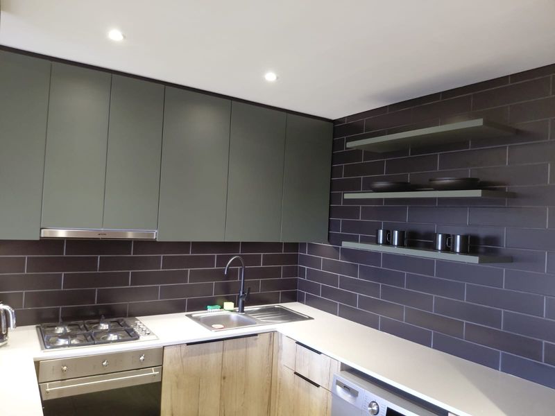 Modern kitchen featuring matte green cabinetry, dark subway tile backsplash, integrated appliances, and floating shelves with recessed lighting.
