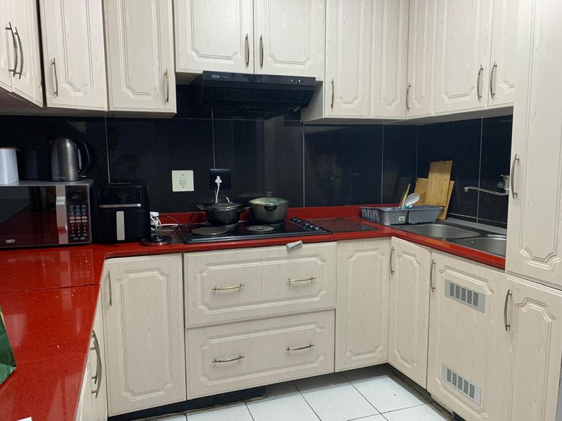 A functional kitchen featuring light-toned cabinetry, a contrasting red stone countertop, black tiled splashback, and integrated hob with extractor fan.
