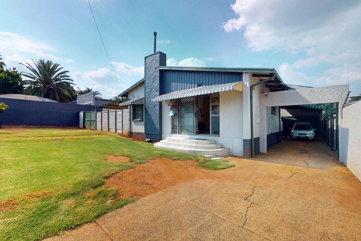Rear view of the property featuring a concrete driveway, carport, and a spacious garden area with perimeter walling and security fencing.