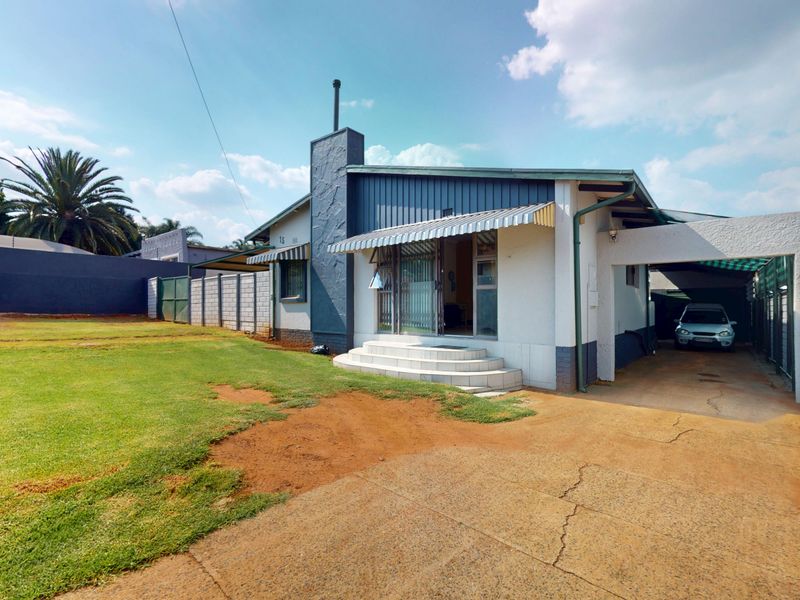 Rear view of the property featuring a concrete driveway, carport, and a spacious garden area with perimeter walling and security fencing.