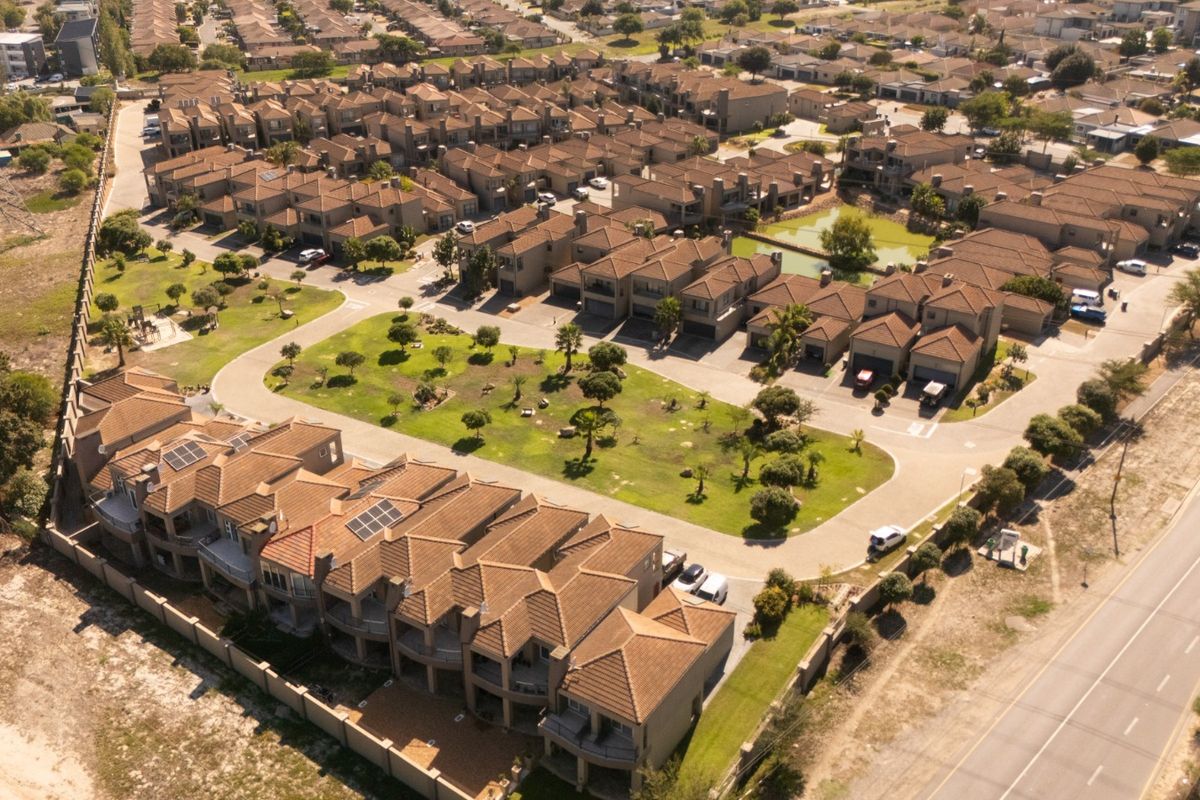 An aerial view of a townhouse complex featuring multiple residential units, tiled roofs, communal green spaces, and paved access roads.