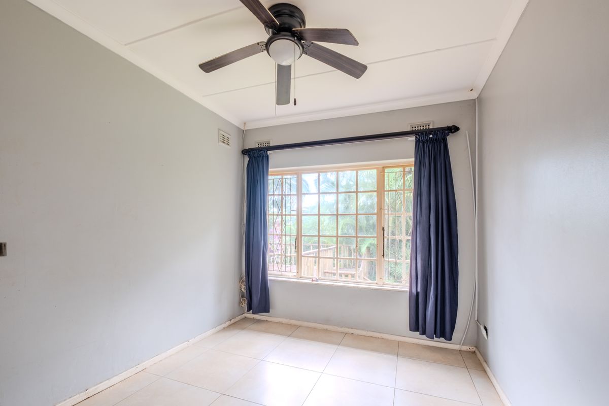 A bright, empty bedroom featuring tiled flooring, a ceiling fan, and large windows fitted with security bars.