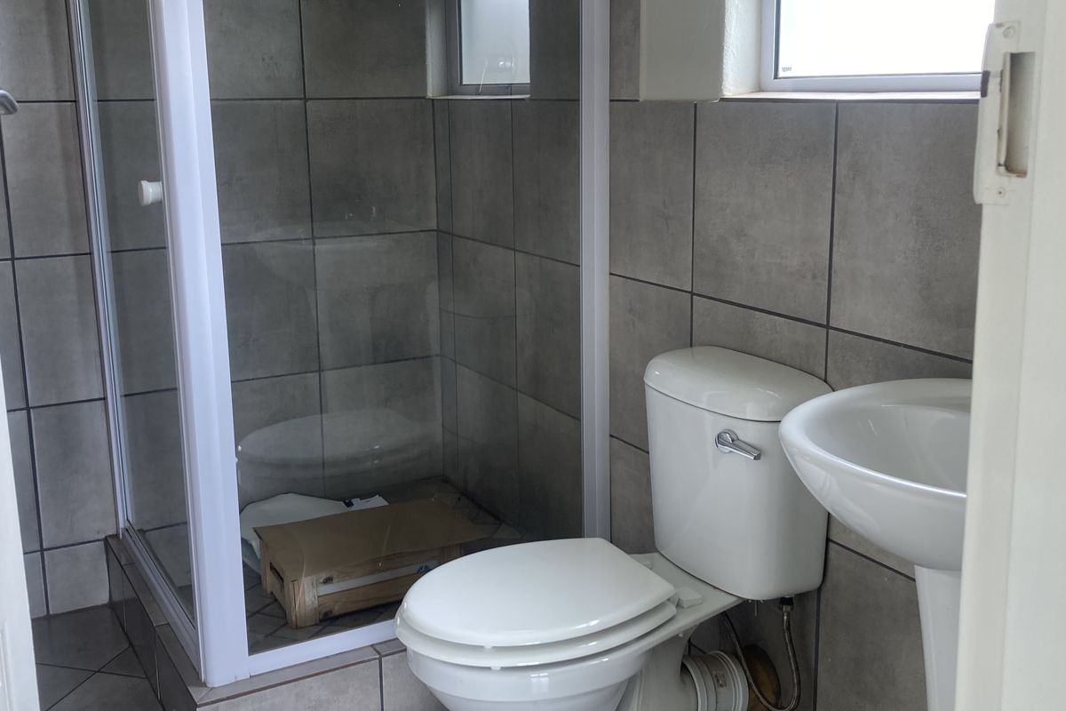 A clean, functional bathroom featuring a glass shower enclosure, pedestal basin, and toilet with neutral grey wall and floor tiling.