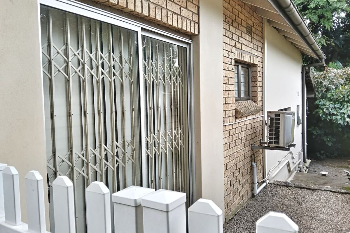 Rear exterior view of the cottage featuring a brick facade, sliding glass door with security gate, and an external air conditioning unit.