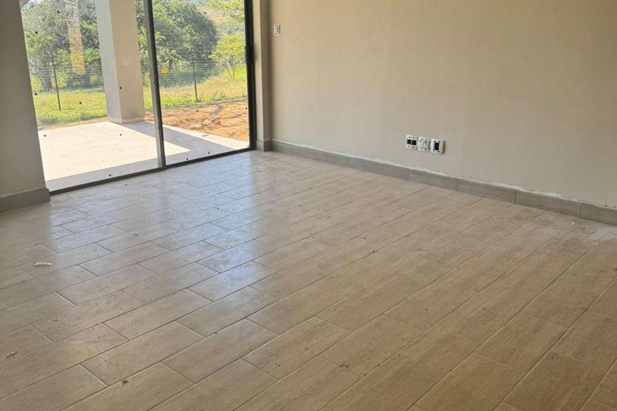 An empty, well-lit living room featuring neutral ceramic tile flooring and a large sliding glass door providing access to an outdoor patio area.