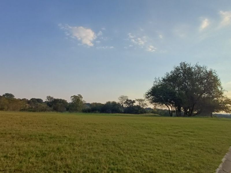 A view of a vacant residential plot featuring a paved road, kerbing, and established trees, offering a clear space for future development.