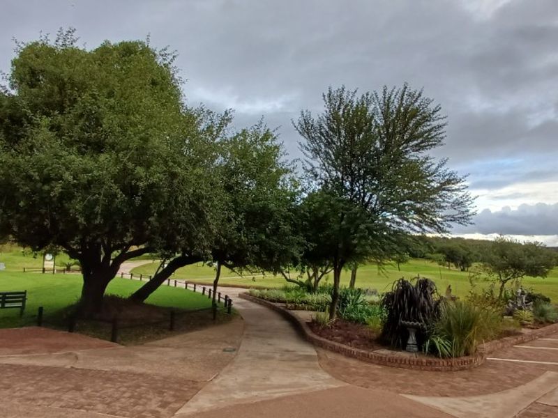 A well-maintained landscaped garden area featuring mature trees, paved pathways, and brick-edged garden beds overlooking an open green space.
