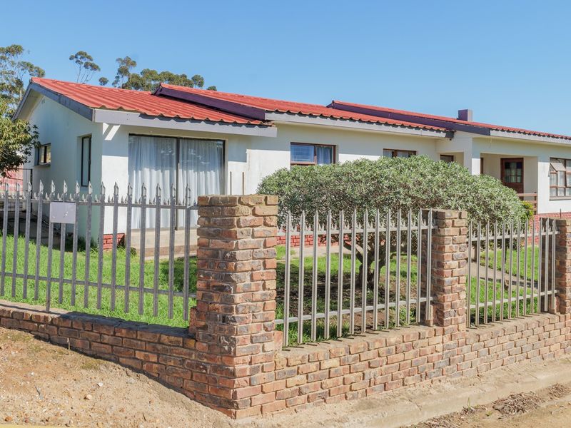 Front view of a single-story house featuring a red corrugated metal roof, brick boundary wall with metal palisade fencing, and a small front garden.