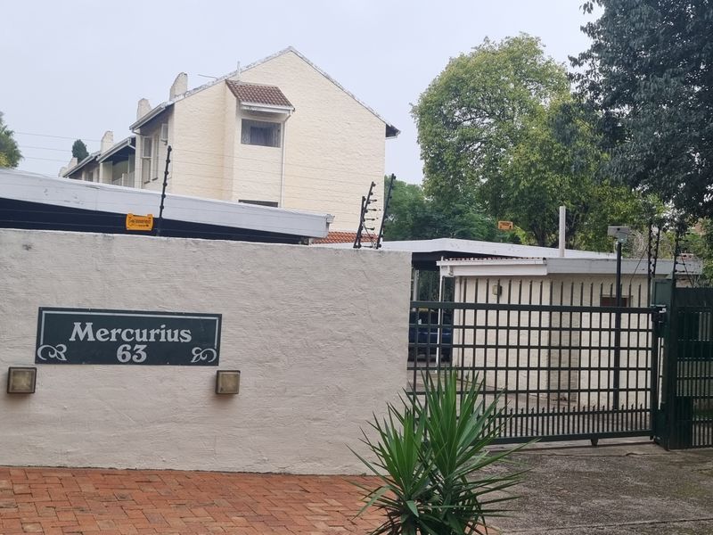Front exterior view of the townhouse complex entrance, featuring a secure gated access point, boundary wall with electric fencing, and paved driveway.
