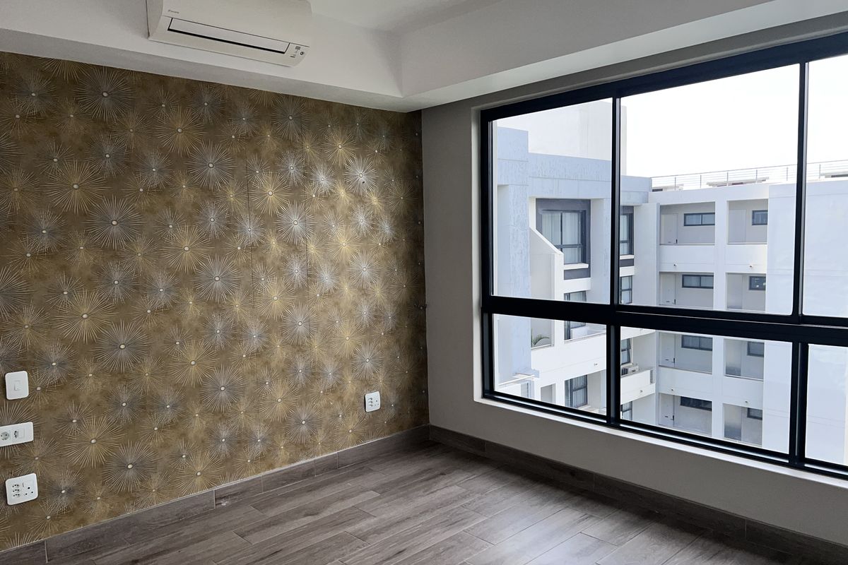 A bright, empty bedroom featuring wood-look tile flooring, a feature wallpaper wall, split-system air conditioning, and large windows overlooking the apartment complex.