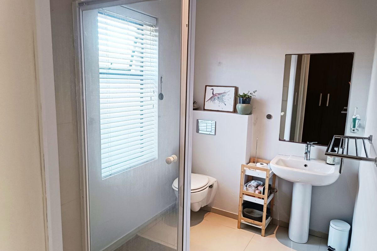 A functional bathroom featuring a pedestal basin, wall-mounted toilet, and a glass-enclosed shower with natural light from a window with venetian blinds.