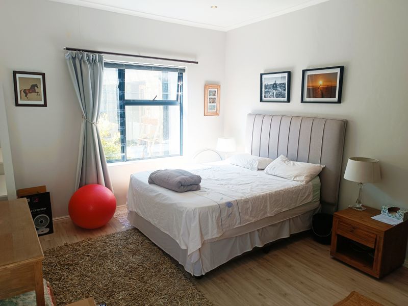 A well-lit bedroom featuring light-toned laminate flooring, neutral walls, and a large window with aluminium frames, providing a clean and functional living space.