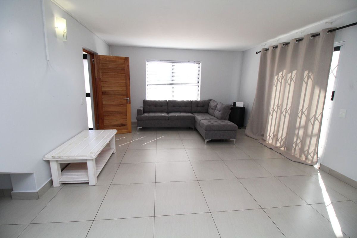 A spacious living room featuring neutral floor tiles, a solid wood door, and large windows allowing for natural light.