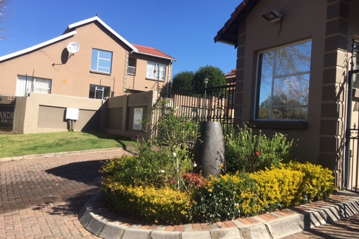 Entrance view of the cluster complex featuring a paved driveway, security gatehouse, perimeter walling with electric fencing, and a landscaped garden bed.