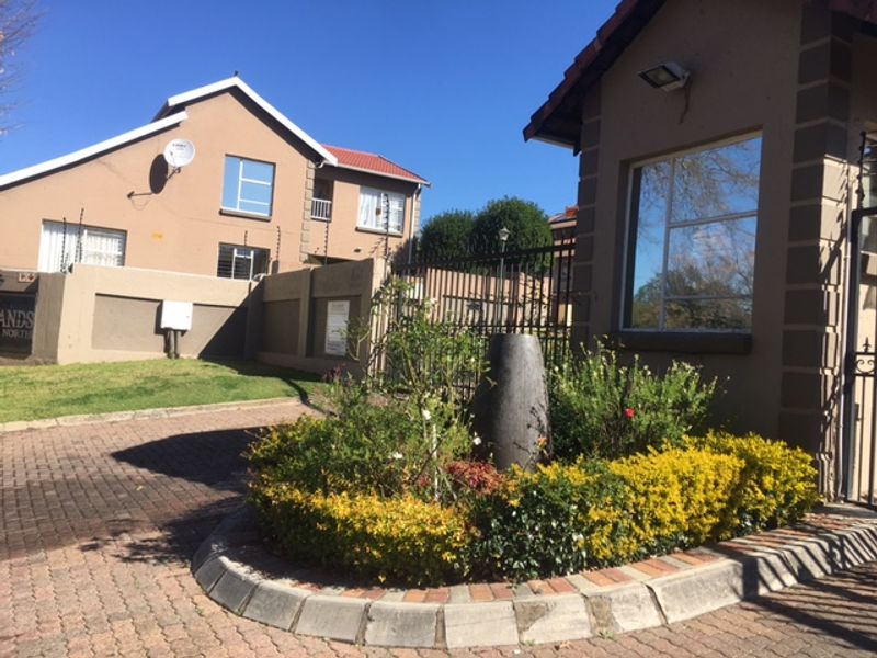 Entrance view of the cluster complex featuring a paved driveway, security gatehouse, perimeter walling with electric fencing, and a landscaped garden bed.