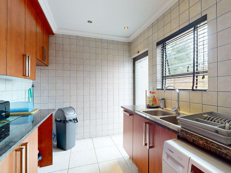 Functional scullery featuring wood-finish cabinetry, granite countertops, a double stainless steel sink, and tiled finishes throughout.