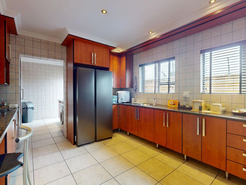 A functional kitchen featuring wood-finish cabinetry, granite countertops, tiled flooring, and a dedicated scullery area visible through the doorway.