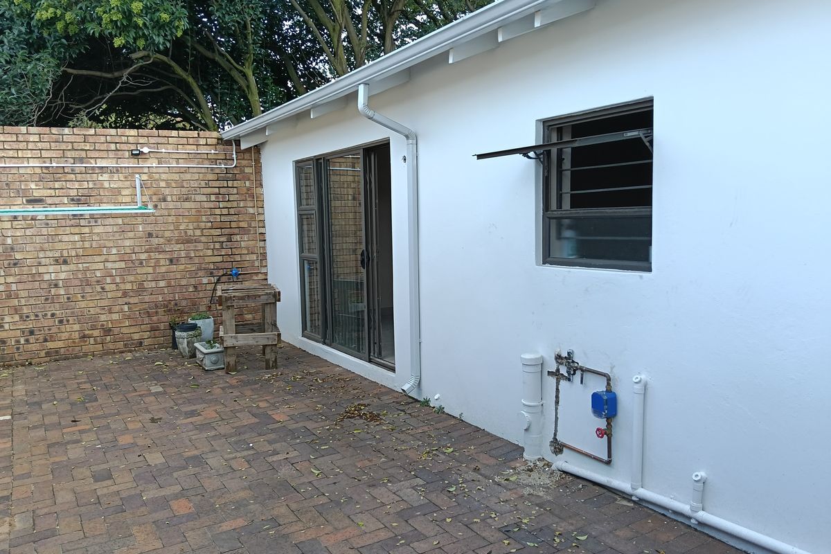 Rear exterior view of the apartment featuring brick paving, a sliding glass door, and exposed exterior plumbing against a white rendered wall.