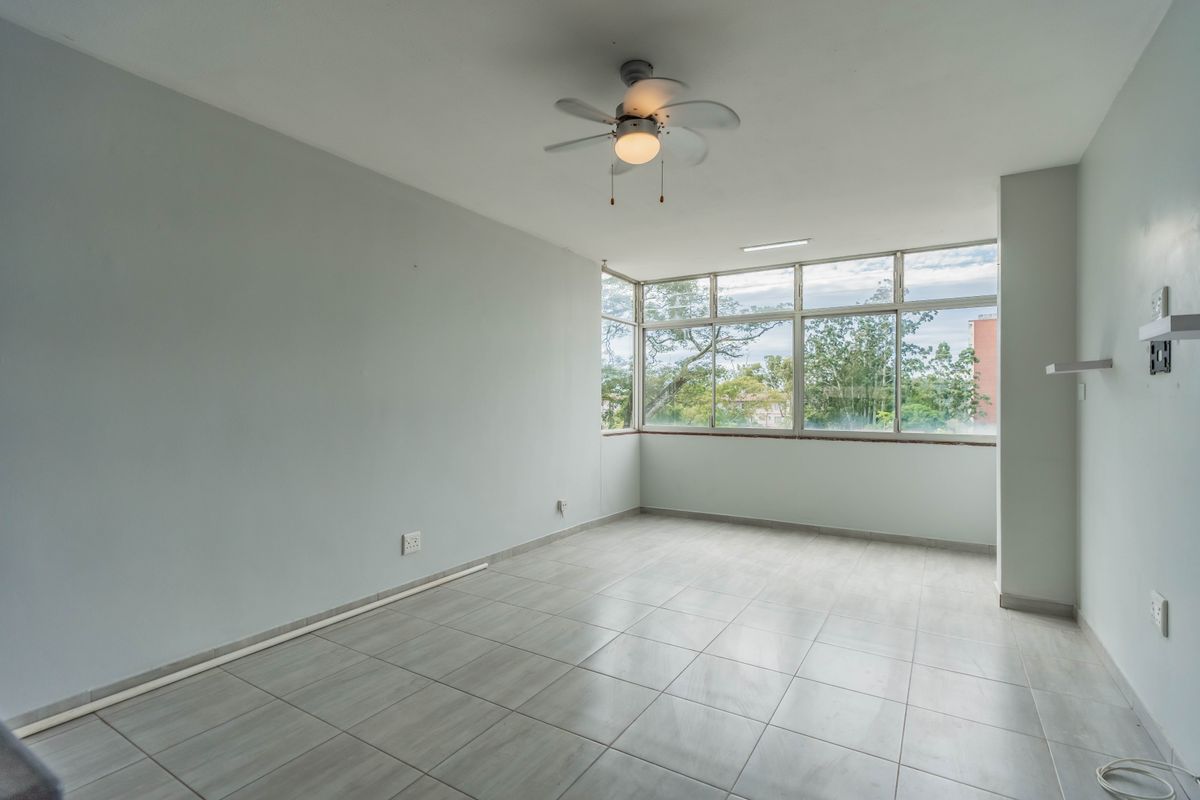 A bright, empty living area featuring large windows, neutral ceramic floor tiles, and a ceiling fan, offering a clean canvas for interior design.