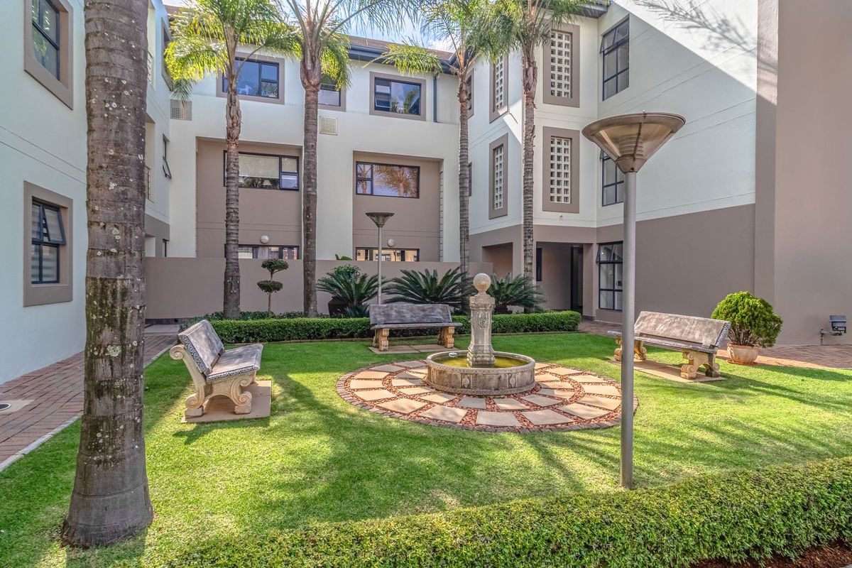 A well-maintained communal garden courtyard featuring a central water fountain, manicured lawn, and paved pathways surrounded by the apartment building facade.