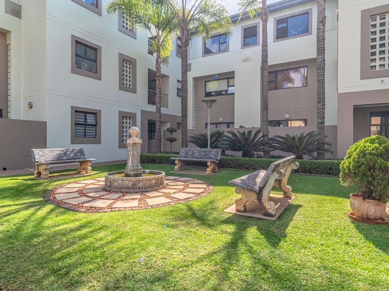 A well-maintained communal garden area featuring a central stone fountain, paved pathways, and established landscaping within an apartment complex courtyard.