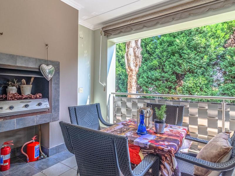 Covered balcony featuring a built-in braai, tiled flooring, and a stainless steel balustrade overlooking lush greenery.