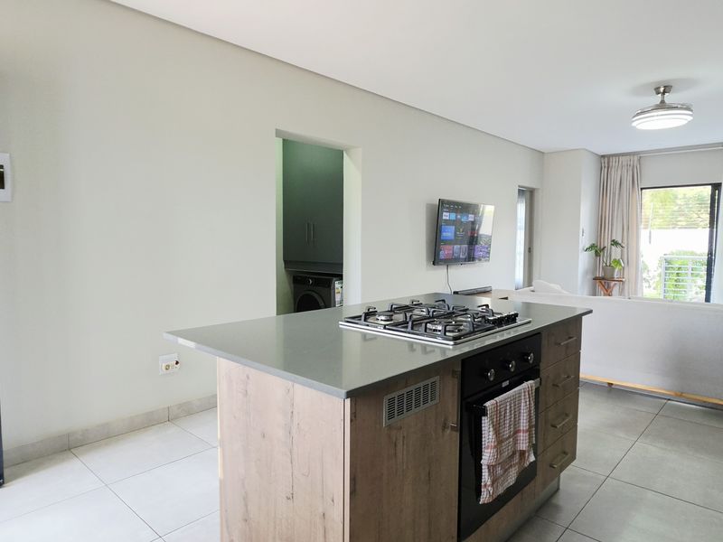 Modern open-plan living area featuring a central kitchen island with integrated gas cooktop and oven, leading to a bright lounge with sliding door access.