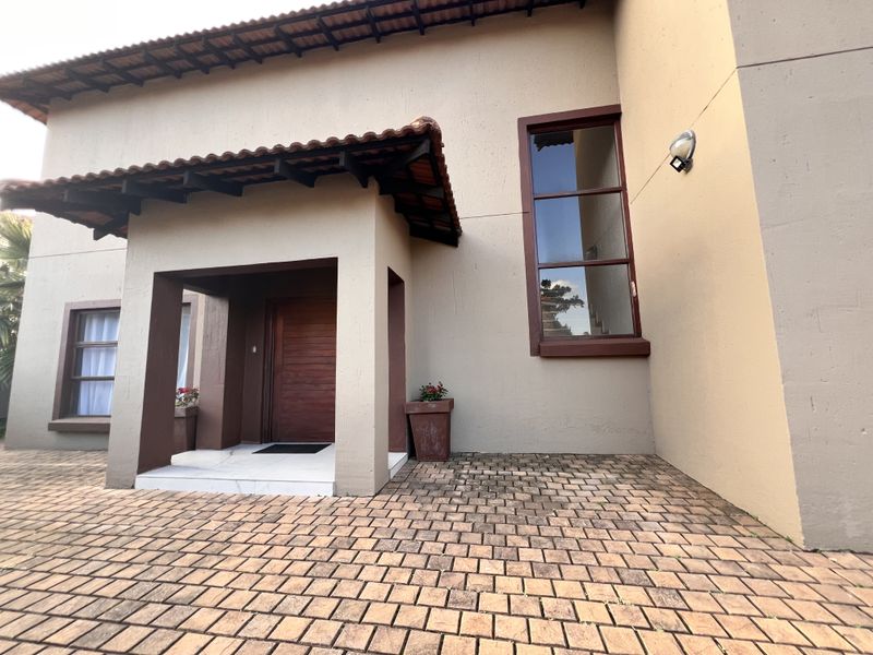 Front entrance of the house featuring a covered porch, wooden door, tall window, and paved driveway area.