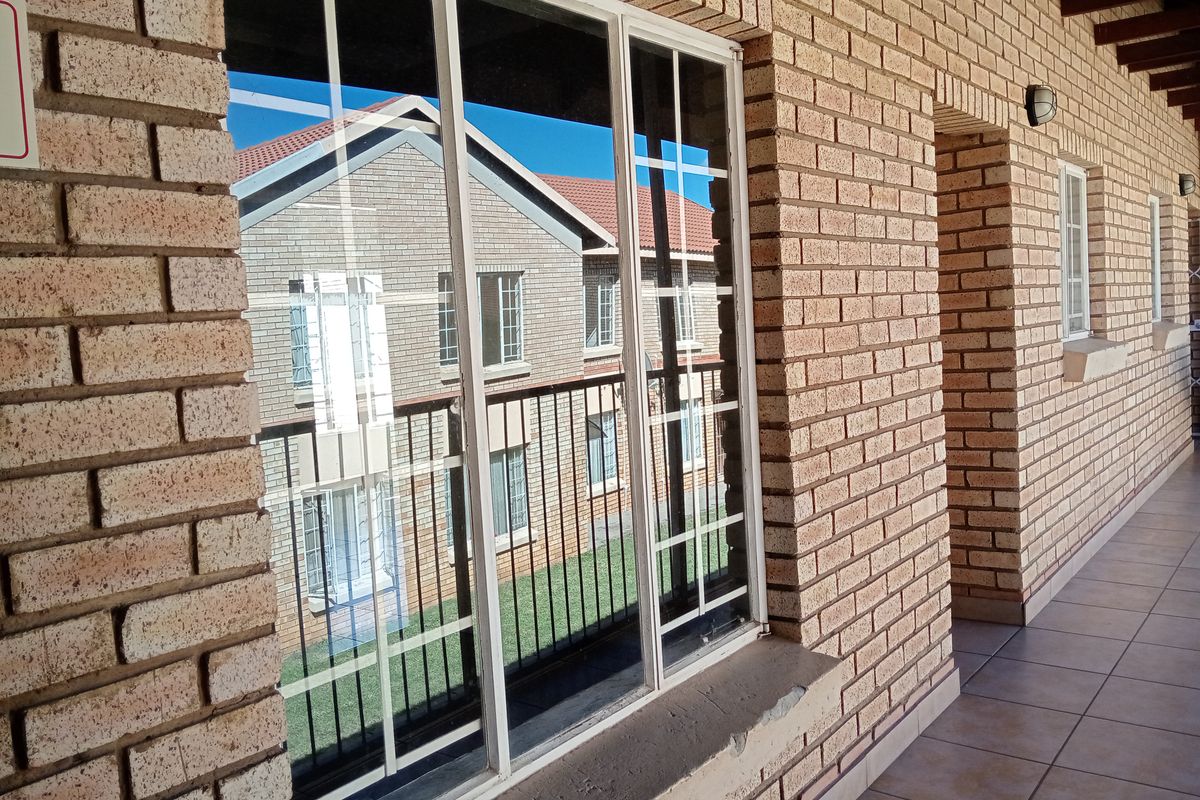 A covered balcony area featuring exposed brick walls, tiled flooring, and a large sliding window with views of the surrounding apartment complex.