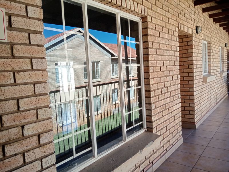 A covered balcony area featuring exposed brick walls, tiled flooring, and a large sliding window with views of the surrounding apartment complex.