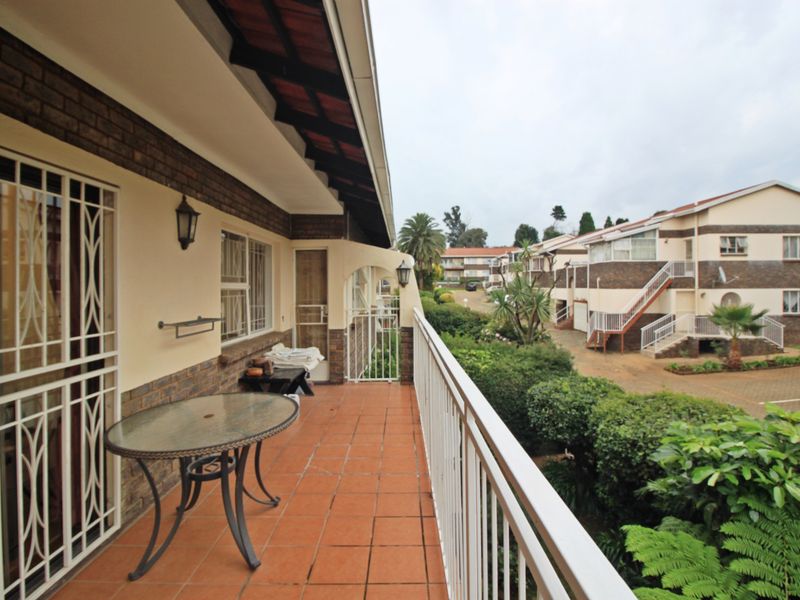 A covered balcony featuring tiled flooring and a metal balustrade, overlooking the complex grounds and surrounding residential units.