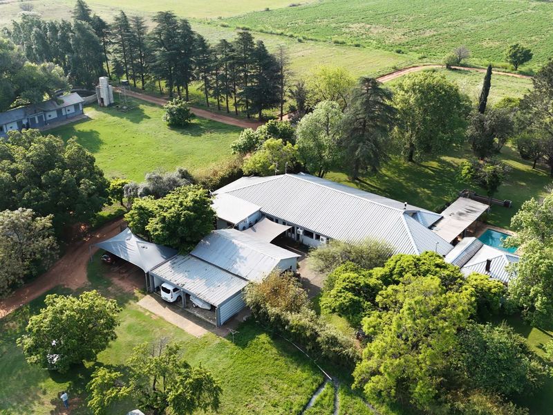 Aerial view of a farm property featuring a main residence with a metal roof, swimming pool, carport, and surrounding outbuildings set within expansive grounds.