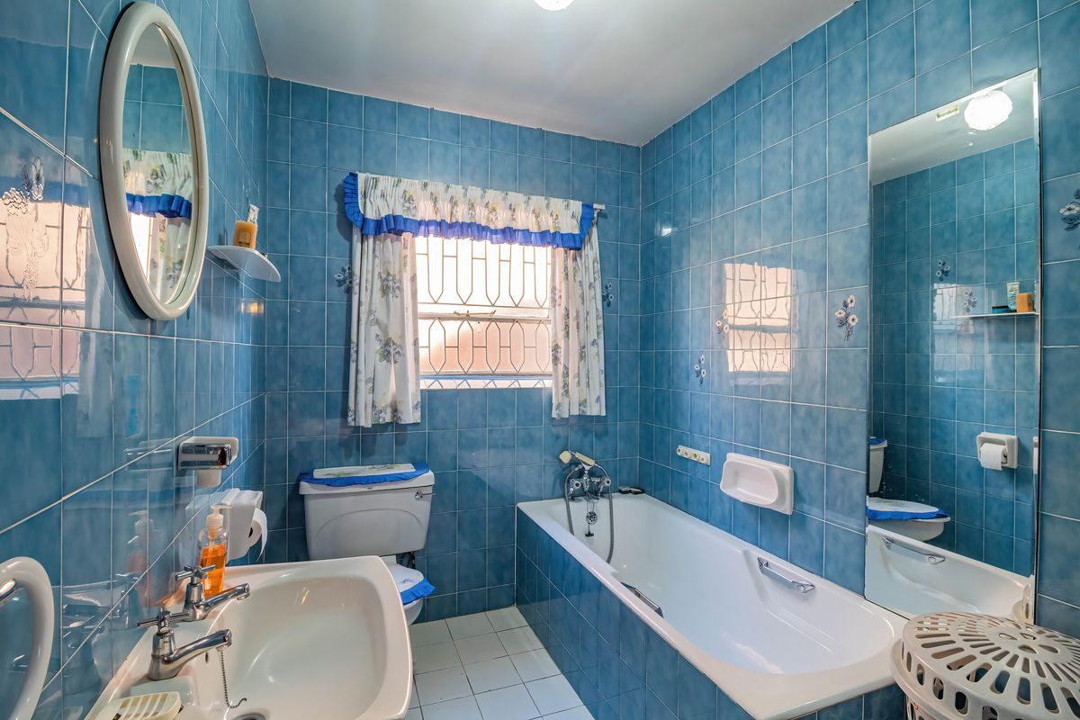 A dated bathroom featuring blue wall tiles, a built-in bathtub, a pedestal sink, and a toilet, with security bars on the window.