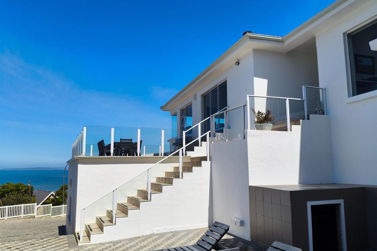 Rear exterior view featuring a raised patio with glass balustrades, external staircase, and paved courtyard area with ocean views.