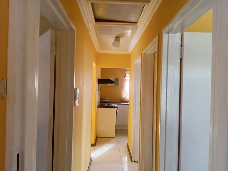 A narrow residential hallway with yellow walls, tiled flooring, and decorative ceiling cornicing, leading towards a kitchen area.