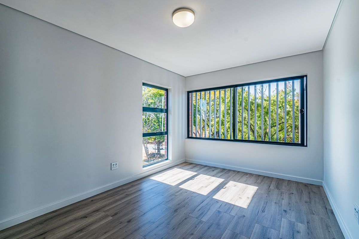 A bright, empty bedroom featuring modern laminate flooring, clean white walls, and large windows with integrated security bars.