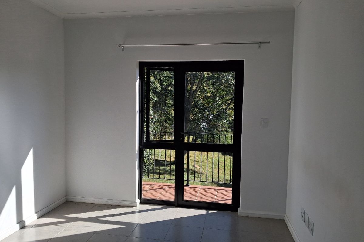 A bright, empty living room featuring neutral tiled flooring and a black aluminium framed glass door leading to an outdoor balcony area.