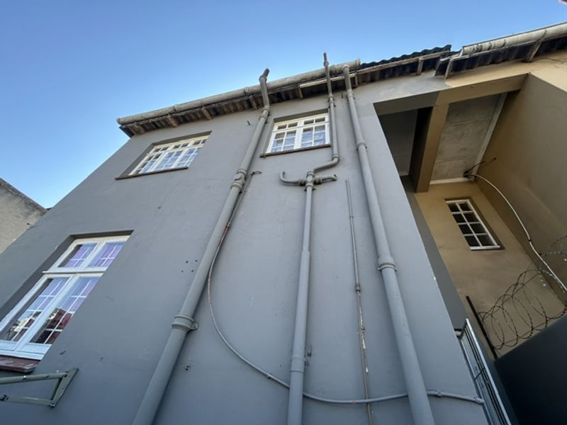 Low-angle view of the rear building facade featuring exposed exterior plumbing, casement windows, and security fencing.