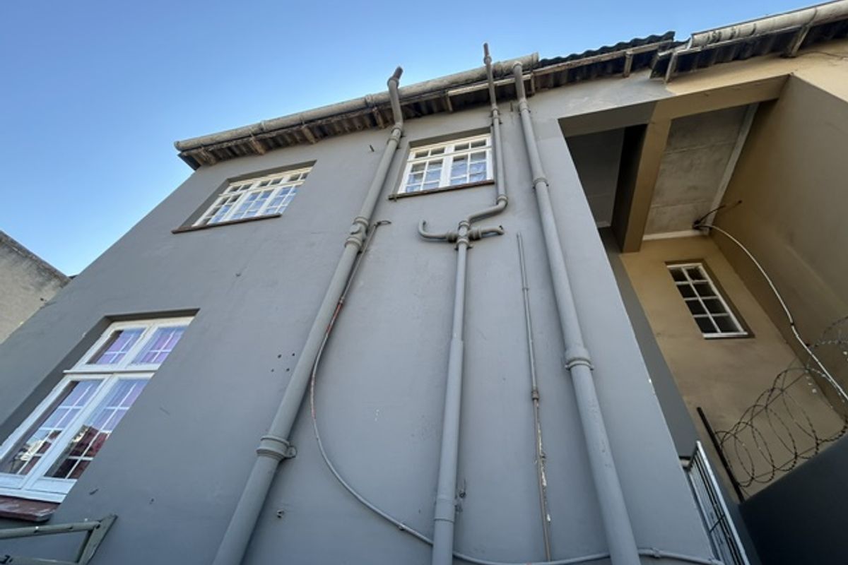 Low-angle view of the rear building facade featuring exposed exterior plumbing, casement windows, and security fencing.