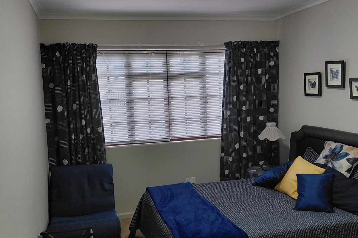 A well-maintained bedroom featuring light-toned laminate flooring, neutral walls, and a window fitted with venetian blinds and curtains.