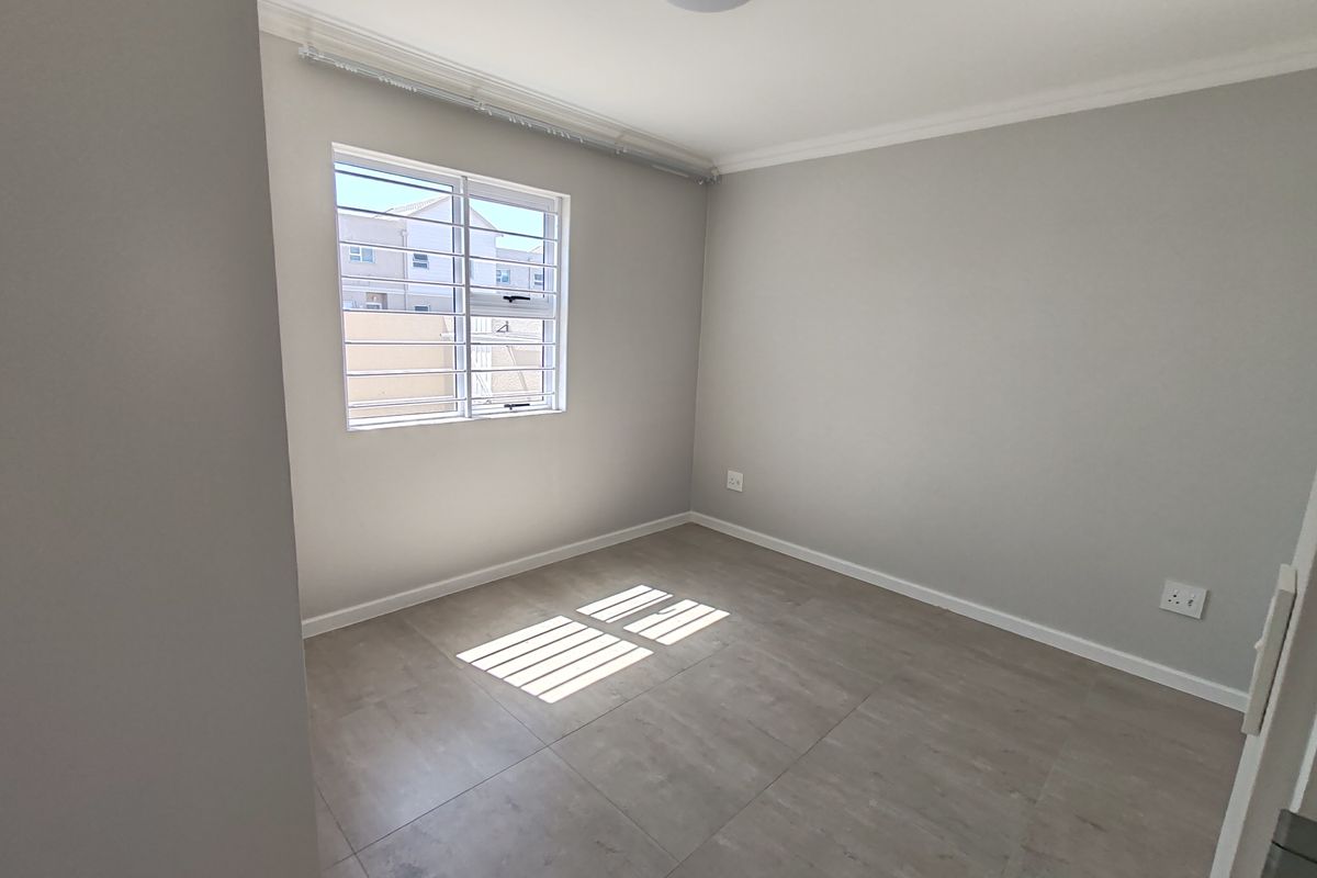 A clean, empty bedroom featuring neutral grey tiled flooring, light-toned walls, and a window fitted with security bars.
