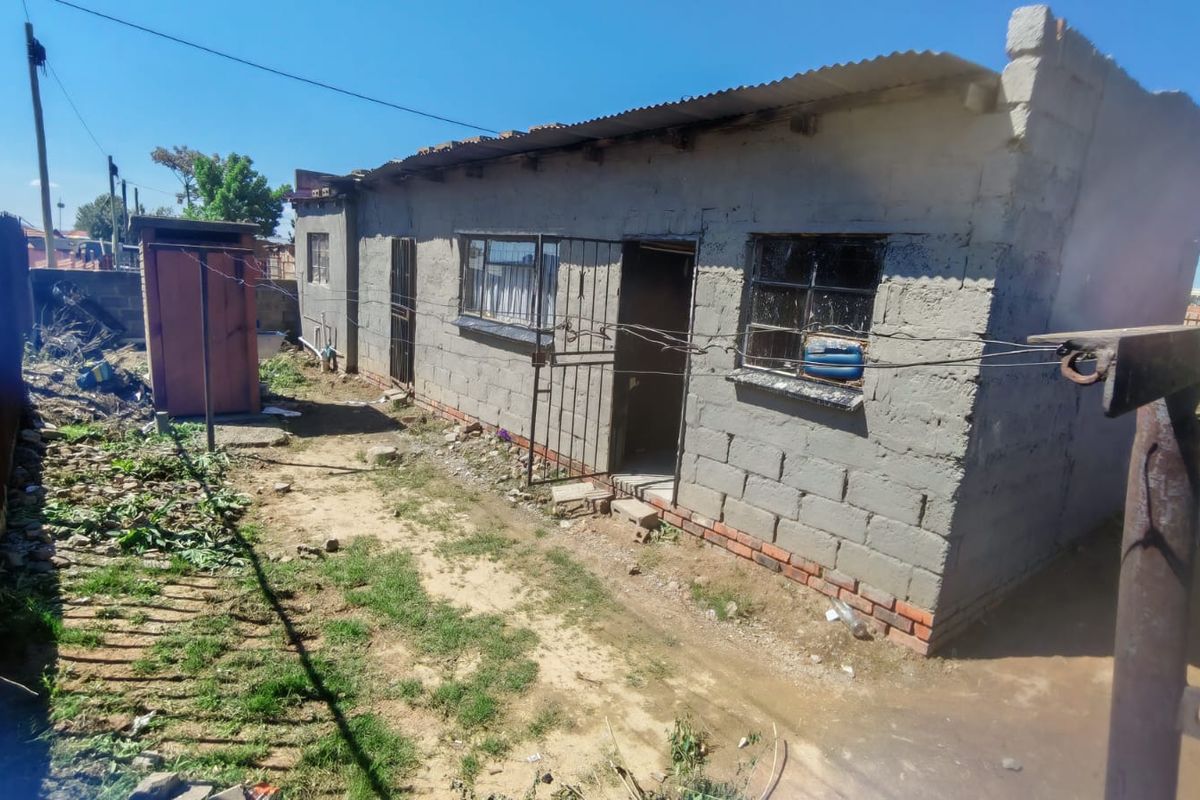 Rear view of a single-story house featuring unplastered concrete block walls, metal security gates, and a corrugated metal roof in need of exterior maintenance.