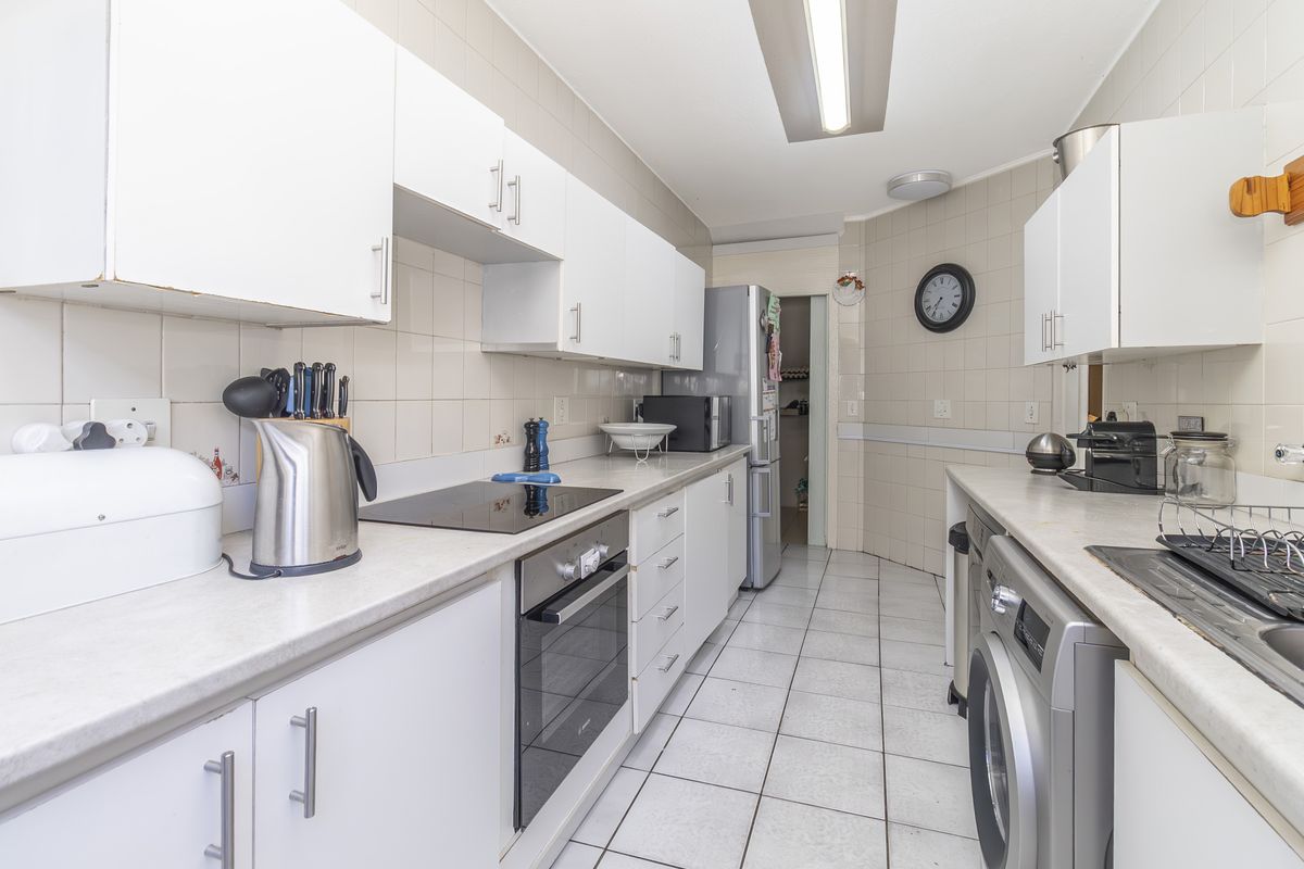 A functional galley-style kitchen featuring white cabinetry, tiled surfaces, built-in oven and hob, and integrated laundry facilities.