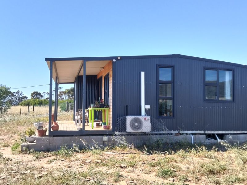 A modern modular home featuring dark corrugated metal cladding, a covered wooden deck, and an external air conditioning unit set on a concrete block foundation.