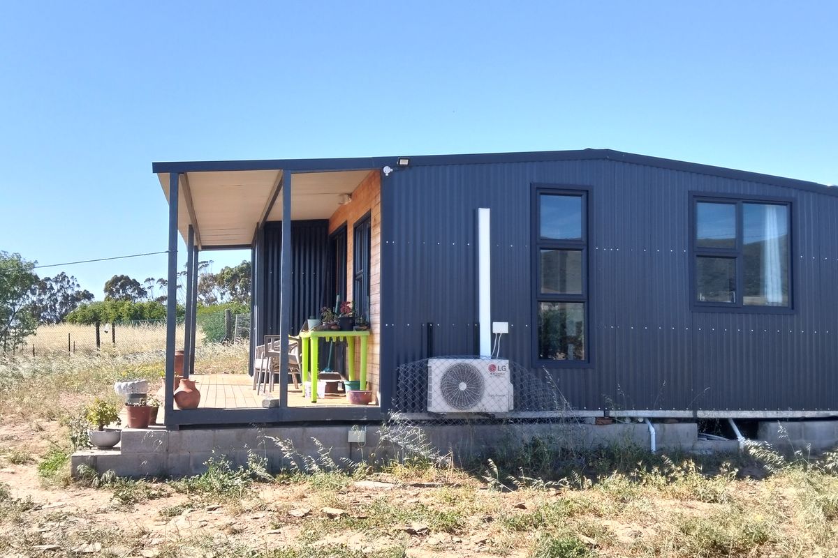 A modern modular home featuring dark corrugated metal cladding, a covered wooden deck, and an external air conditioning unit set on a concrete block foundation.