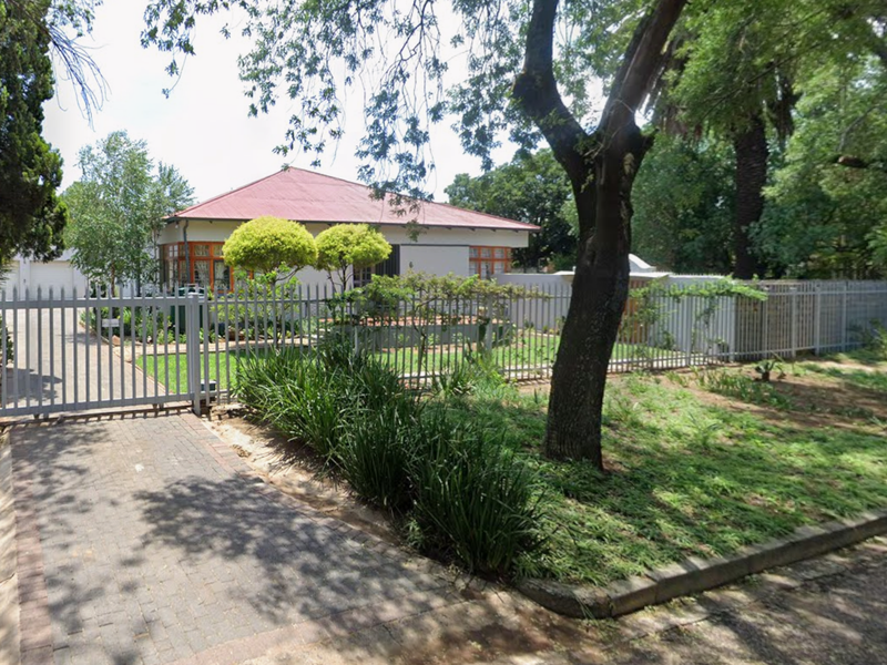 Front view of the property featuring a paved driveway, metal perimeter fencing, and a pitched roof house set within a landscaped garden.