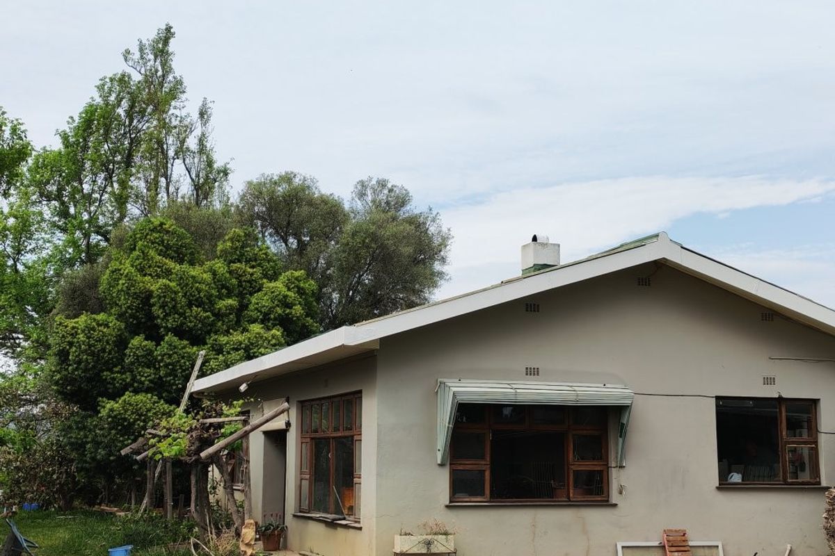Rear view of a farm-style dwelling featuring plastered walls, a pitched roof, chimney, and timber-framed windows with metal awnings set in a garden.