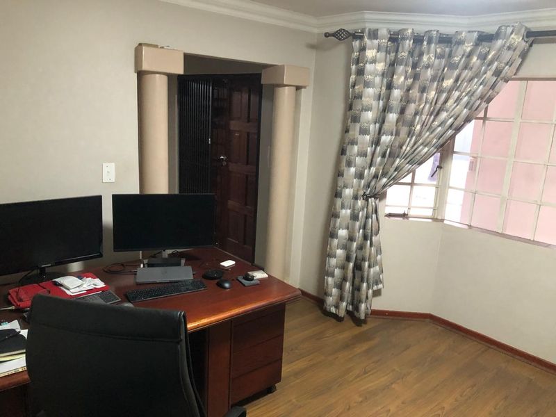 A functional home office space featuring laminate flooring, decorative columns, and a window with security bars.